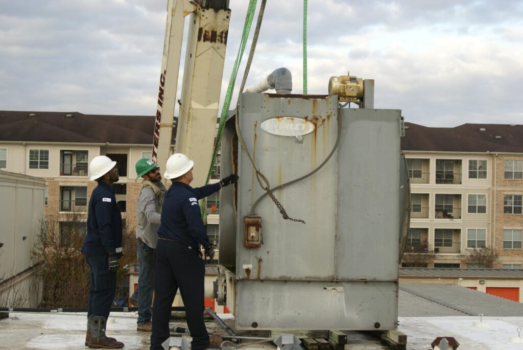 Commercial HVAC installation team using crane to position air conditioning unit at multi-story facility
